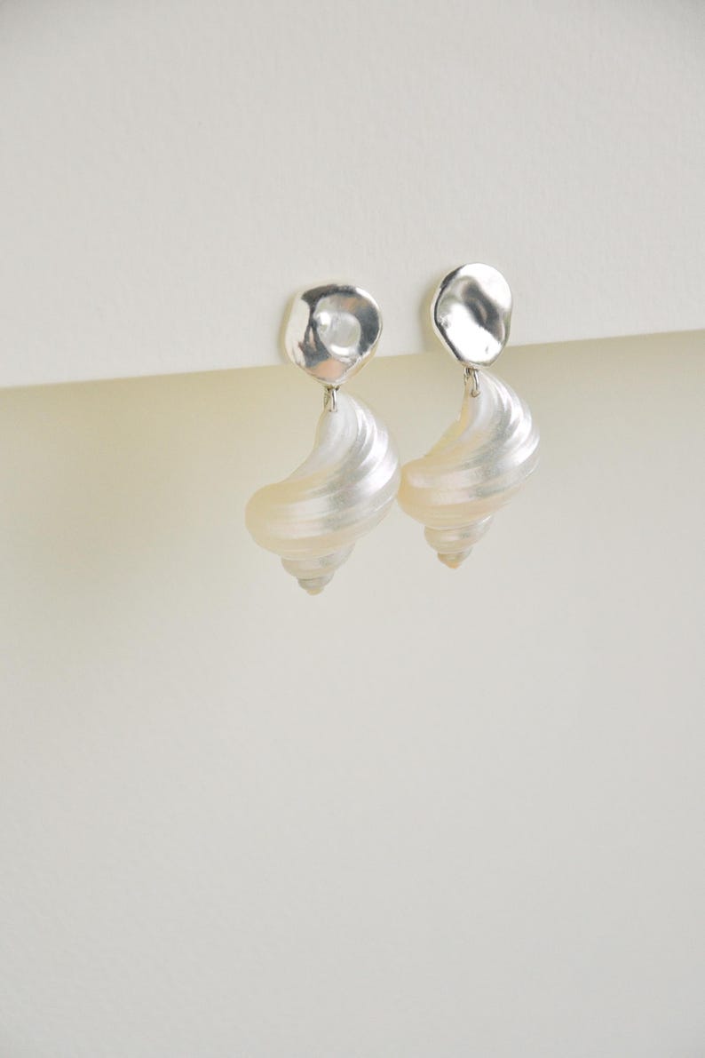 Pu&ograve; includere: Un paio di orecchini eleganti con un perno d'argento di forma irregolare e una goccia perlata a forma di conchiglia. Gli orecchini hanno un design a spirale e una finitura lucida e iridescente.