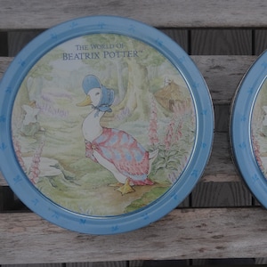 May include: Two blue and white round tin containers with a picture of a duck wearing a blue bonnet and a red and white dress. The text "The World of Beatrix Potter" is printed on the front of each tin.