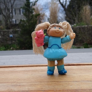 May include: A Cabbage Patch Kid doll with blonde hair in pigtails, wearing a blue dress and holding a pink teddy bear.