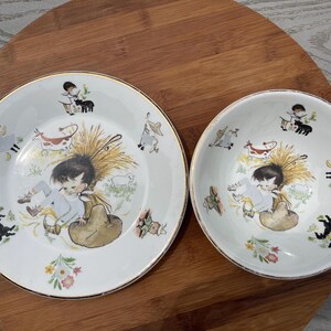May include: A white ceramic plate and bowl set featuring a gold rim and a whimsical illustration of a child sitting in a haystack surrounded by small farm animals, including a cow, a goat, and a black cat, with colorful floral accents.