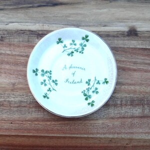 May include: A white ceramic souvenir plate with a gold rim, featuring green shamrock sprigs and the text "A souvenir of Ireland" in a delicate script. The plate is round and rests on a wooden surface.