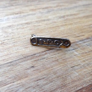 May include: A small, antique-style pin with the letters "E.F.C." engraved on it. The pin is a bronze color and has a simple, rectangular shape with rounded ends. It is set against a wooden background.