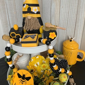 May include: A tiered tray decorated with a bee theme. The tray features a bee gnome, honeycomb blocks, a wooden honey dipper, a yellow mug with the text "Busy Bee", and a wooden sign that says "bee kind". The tray is decorated with yellow and black beads, yellow and black fabric hearts, and yellow flowers.