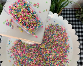 Op de afbeelding: Een witte taartstandaard met twee lagen, gevuld met kleurrijke sprinkels. De sprinkels zijn een mix van roze, groen, blauw, geel en goud. Een doorzichtige plastic schep is gevuld met sprinkels en staat op de bovenste laag.