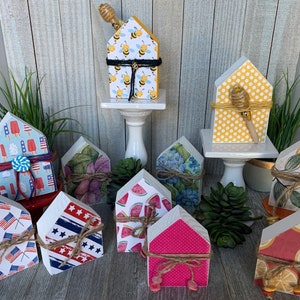 May include: A collection of 10 wooden houses decorated with various summer-themed patterns. The houses are painted in shades of blue, red, yellow, green, and pink. Each house features a different design, including popsicles, watermelon, bees, lemons, and flowers. The houses are arranged on a wooden surface with a white background.
