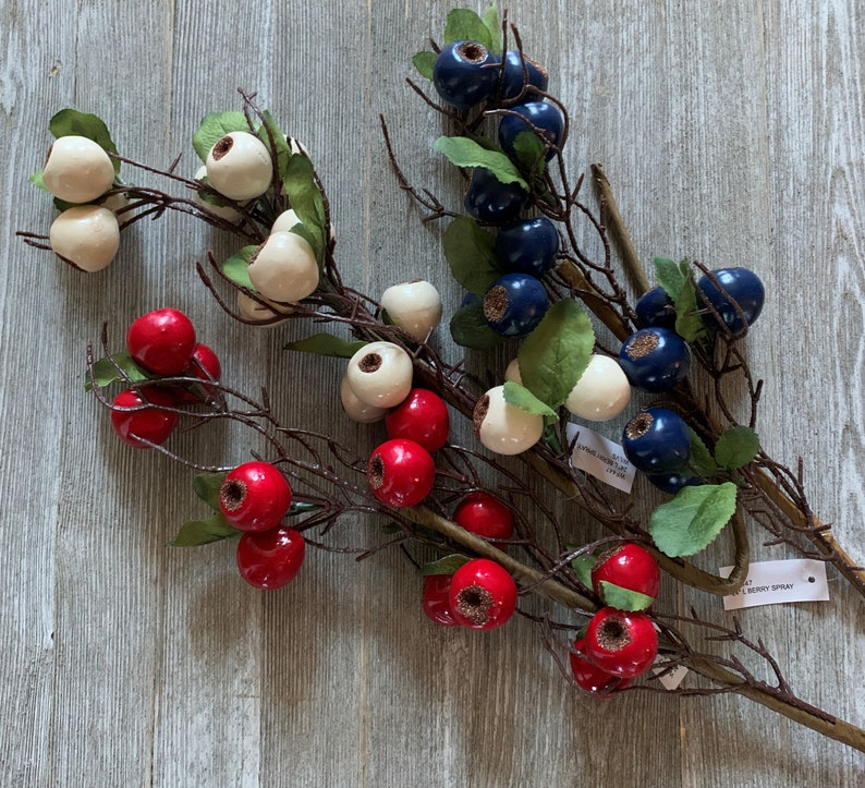 May include: Three sprigs of artificial berries with white, red, and blue berries. The sprigs are arranged on a wooden surface. The text "Berry Spray" is visible on the bottom right of the image.