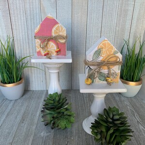 K&ouml;nnte beinhalten: Zwei kleine Holzh&auml;user, die mit Zitronen- und Blumenmustern verziert sind. Die H&auml;user stehen auf wei&szlig;en Sockeln mit kleinen gr&uuml;nen Sukkulenten.