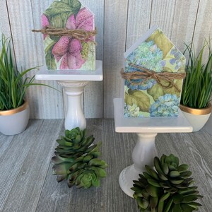 K&ouml;nnte beinhalten: Zwei kleine Holzh&auml;user, die mit Blumenmustern verziert sind. Das Haus links hat rosa Blumen und gr&uuml;ne Bl&auml;tter, w&auml;hrend das Haus rechts blaue Blumen und gr&uuml;ne Bl&auml;tter hat. Beide H&auml;user stehen auf wei&szlig;en Sockeln mit gr&uuml;nen Sukkulenten.