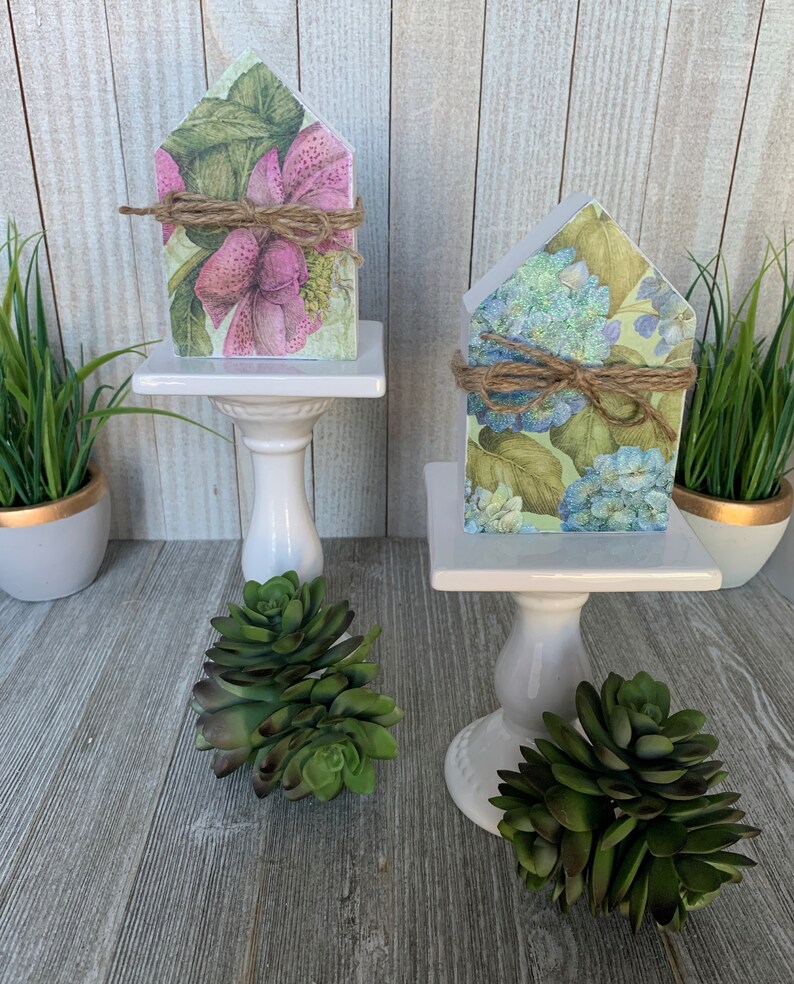K&ouml;nnte beinhalten: Zwei kleine Holzh&auml;user, die mit Blumenmustern verziert sind. Das Haus links hat rosa Blumen und gr&uuml;ne Bl&auml;tter, w&auml;hrend das Haus rechts blaue Blumen und gr&uuml;ne Bl&auml;tter hat. Beide H&auml;user stehen auf wei&szlig;en Sockeln mit gr&uuml;nen Sukkulenten.