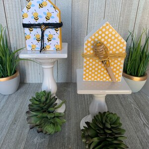 K&ouml;nnte beinhalten: Zwei Holzh&auml;user, die mit einem Bienen- und Punkte-Muster verziert sind. Die H&auml;user stehen auf wei&szlig;en Sockeln mit kleinen gr&uuml;nen Sukkulenten davor.