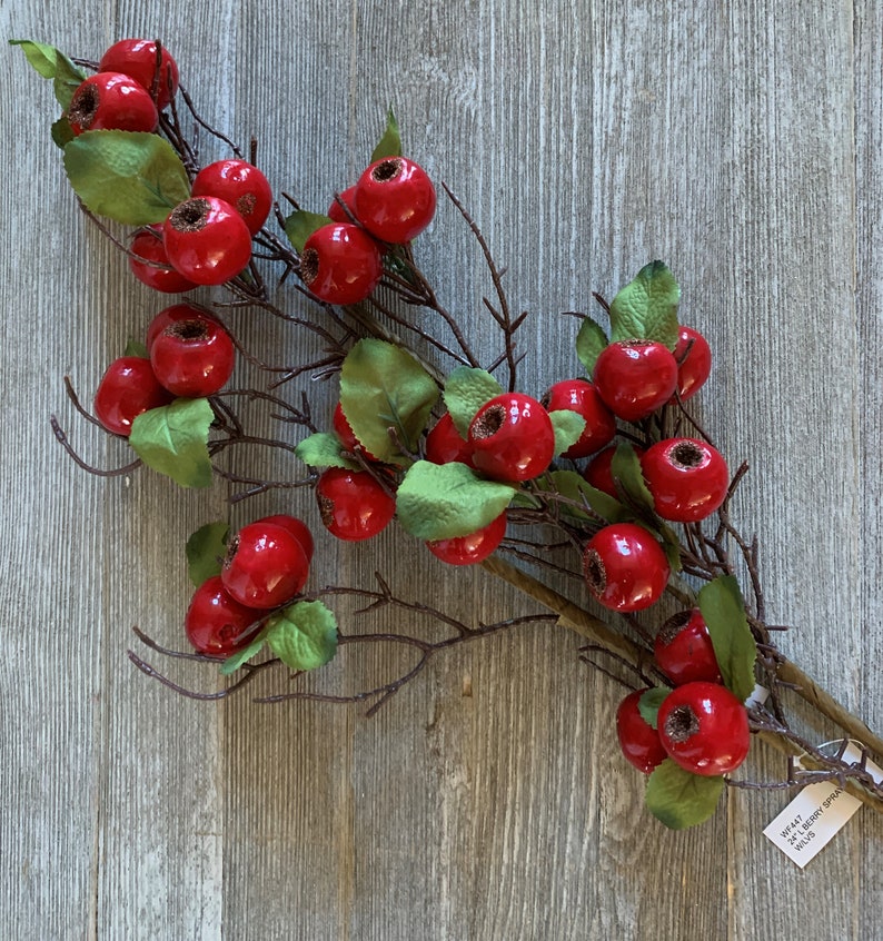 May include: A sprig of artificial red berries with green leaves on a brown wooden background. The sprig is about 30 inches long and has a tag that says "WF442 Red Berry Sprig 30"