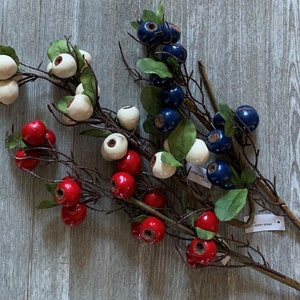 May include: Three sprigs of artificial berries with red, white, and blue berries. The sprigs are on a gray wooden surface. The text "berry spray" is visible on one of the sprigs.