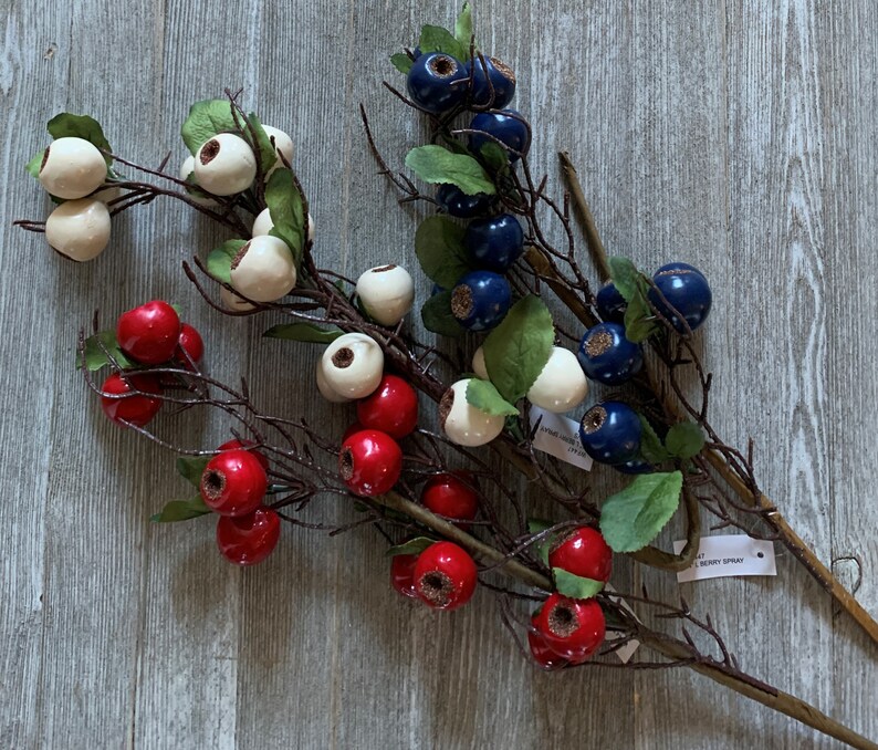May include: Three sprigs of artificial berries with red, white, and blue berries. The sprigs are on a gray wooden surface. The text "berry spray" is visible on one of the sprigs.