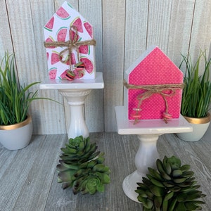 K&ouml;nnte beinhalten: Zwei kleine Holzh&auml;user, die mit Sommermotiven verziert sind. Ein Haus ist pink mit wei&szlig;en Punkten und das andere ist wei&szlig; mit Wassermelonenmuster. Beide H&auml;user haben eine braune Kordelschleife und stehen auf wei&szlig;en Sockeln.