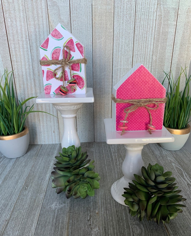 K&ouml;nnte beinhalten: Zwei kleine Holzh&auml;user, die mit Sommermotiven verziert sind. Ein Haus ist pink mit wei&szlig;en Punkten und das andere ist wei&szlig; mit Wassermelonenmuster. Beide H&auml;user haben eine braune Kordelschleife und stehen auf wei&szlig;en Sockeln.