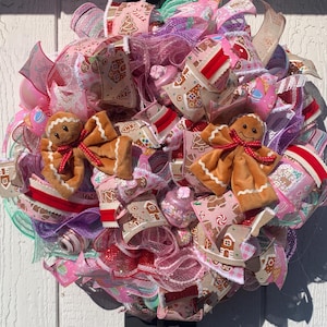 May include: A festive Christmas wreath with a gingerbread theme. The wreath features plush gingerbread men, pink and red ribbons, and decorative elements. The design incorporates various patterns, including gingerbread houses and candy canes, creating a cheerful holiday decoration.