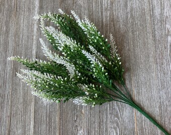 May include: A bundle of artificial greenery with white and green sprigs. The faux plant has a green stem and is arranged on a wooden surface. The sprigs have small white buds and are a mix of green and white.