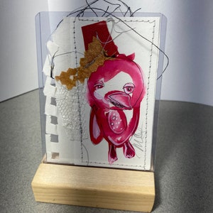Peut inclure: Une illustration d'oiseau rose et blanc avec des accents rouges est encadrée dans un cadre en plastique transparent et montée sur un support en bois. L'oiseau est entouré de morceaux de tissu et de papier.