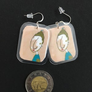 Peut inclure: Une paire de boucles d'oreilles avec une illustration de dessin animé d'une personne avec un chapeau vert, une peau blanche et des vêtements bleus. Les boucles d'oreilles sont en plastique transparent et ont des crochets argentés.
