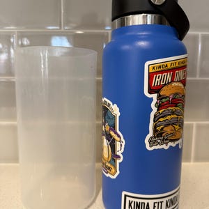 Puede incluir: Una botella de agua azul con tapa y asa negras, y un vaso de plástico transparente. La botella tiene pegatinas, incluyendo "IRON DINER" y "Kinda Fit Kinda Fat".
