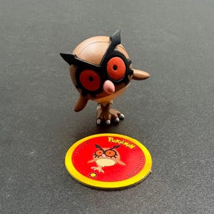 May include: A brown and tan Pokemon figure of an owl-like creature with large red eyes and black markings. A red and yellow Pokemon disc is in front of the figure, with the Pokemon logo and a smaller image of the owl.
