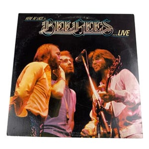 Puede incluir: Una portada de disco de vinilo con una actuación en directo de la banda The Bee Gees. La portada es negra con el nombre de la banda en letras blancas y las palabras "Here at Last...Live" en letras blancas. La imagen muestra a tres hombres en el escenario, dos con micrófonos y uno con una guitarra.