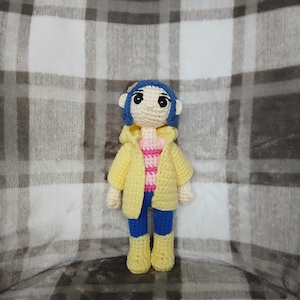 May include: A handmade crochet doll featuring blue hair, a yellow jacket, a pink and white striped shirt, blue pants, and yellow boots. The doll is crafted with a soft, textured yarn and has a whimsical, cartoonish appearance. The doll is standing on a plaid background.