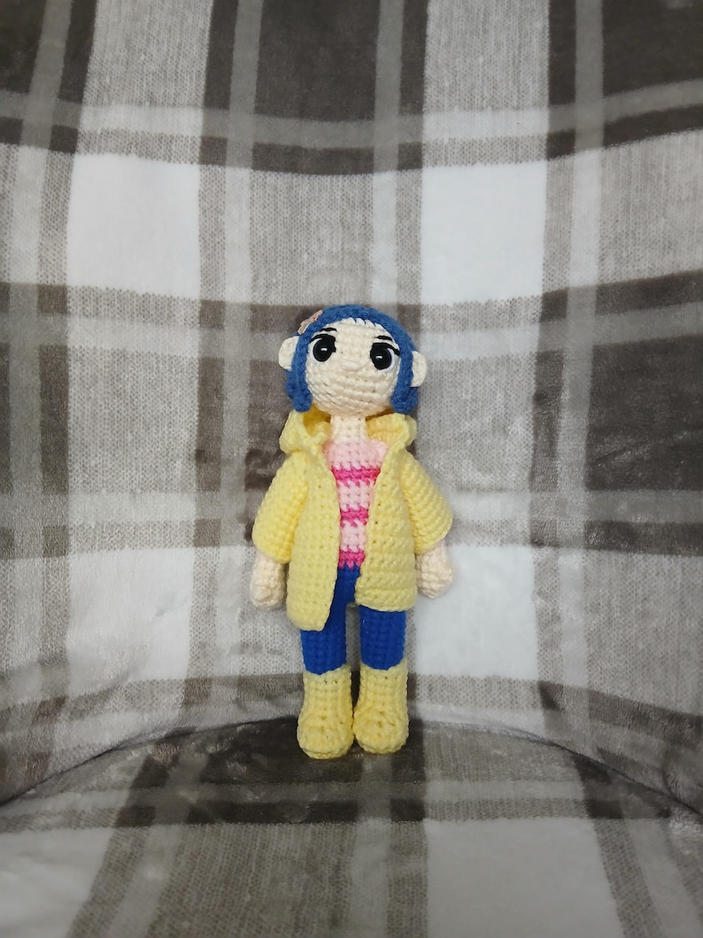 May include: A handmade crochet doll featuring blue hair, a yellow jacket, a pink and white striped shirt, blue pants, and yellow boots. The doll is crafted with a soft, textured yarn and has a whimsical, cartoonish appearance. The doll is standing on a plaid background.