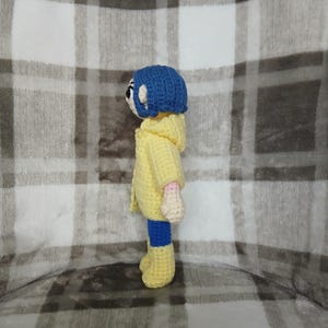 May include: A handmade crochet doll featuring a blue-haired character wearing a yellow hooded jacket, blue pants, and yellow boots. The doll has a white face with black eyes and pink hands. The doll is approximately 6 inches tall.
