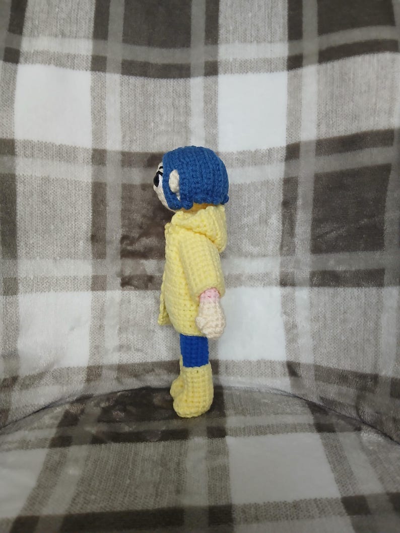 May include: A handmade crochet doll featuring a blue-haired character wearing a yellow hooded jacket, blue pants, and yellow boots. The doll has a white face with black eyes and pink hands. The doll is approximately 6 inches tall.
