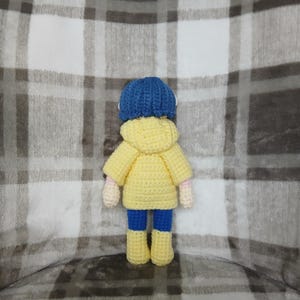 May include: A handmade crochet doll wearing a blue hat, yellow jacket, blue pants, and yellow boots. The doll has pink hands and is standing on a plaid blanket. The doll is a unique, handcrafted item.