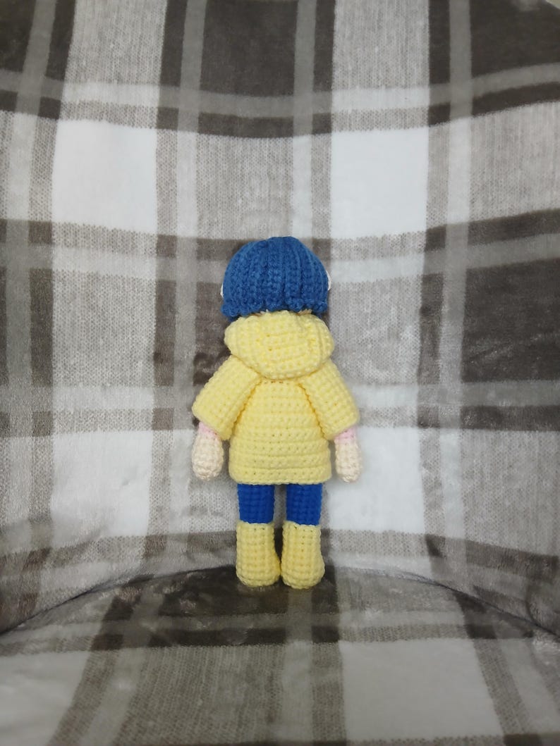 May include: A handmade crochet doll wearing a blue hat, yellow jacket, blue pants, and yellow boots. The doll has pink hands and is standing on a plaid blanket. The doll is a unique, handcrafted item.