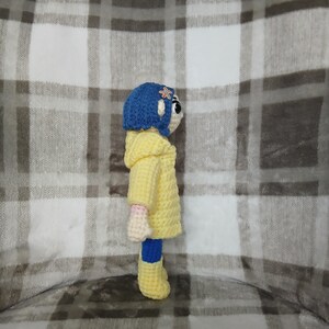 May include: A crocheted doll of a character from a movie, wearing a yellow coat, blue boots, and a blue hat with a flower. The doll is handmade with a white face and pink arms. The background is a plaid pattern.