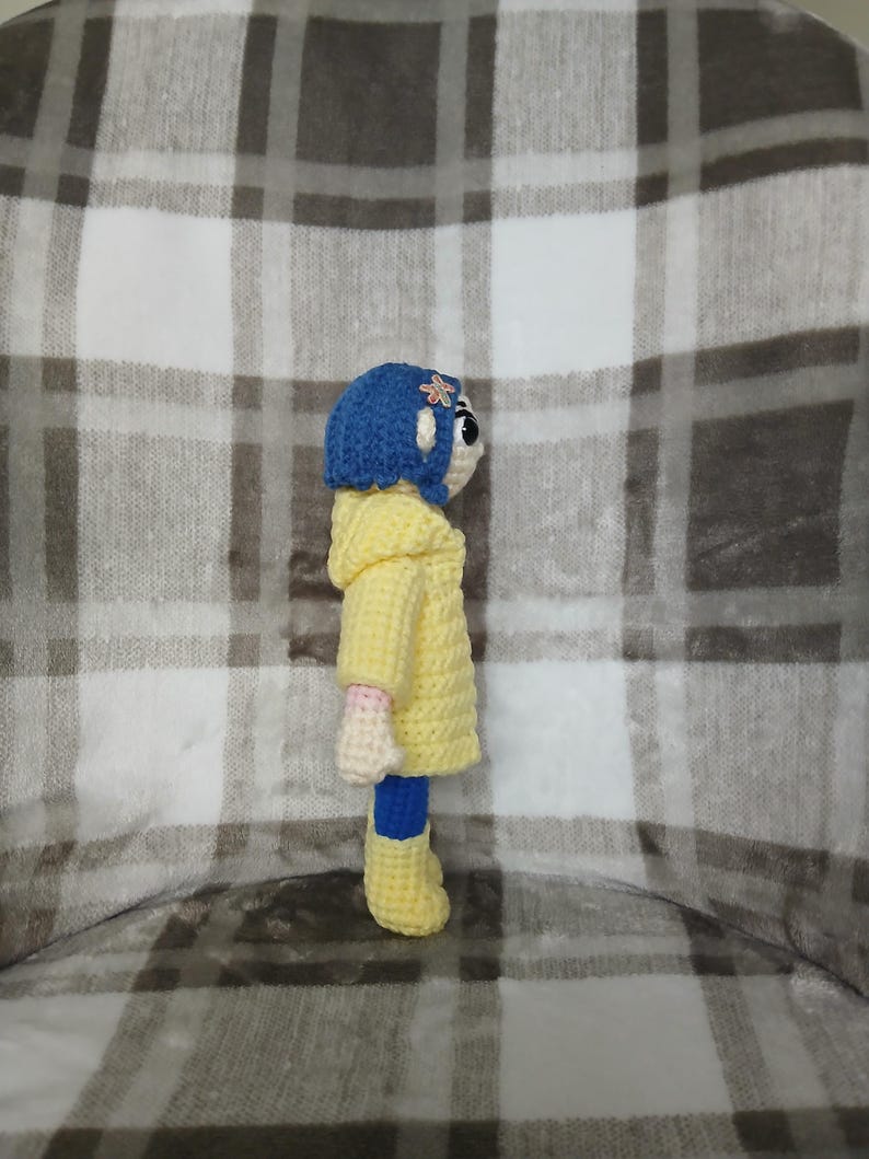 May include: A crocheted doll of a character from a movie, wearing a yellow coat, blue boots, and a blue hat with a flower. The doll is handmade with a white face and pink arms. The background is a plaid pattern.