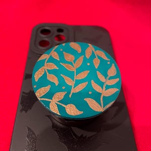 May include: A teal blue phone pop socket with a gold leaf design. The pop socket is on a black phone case with a bat pattern.