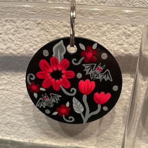 May include: A black circular keychain with a painted design of red flowers, gray leaves, and bats. The bats have red eyes and are flying.