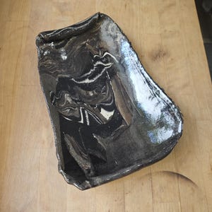 Könnte beinhalten: Ein rechteckiges Keramiktablett mit unregelmäßigen, handgeformten Kanten und einer glänzenden Oberfläche. Das Design zeigt ein marmoriertes Muster aus schwarzen, anthrazitfarbenen und weißen Wirbeln, das ein fließendes, kontrastreiches Bild ergibt.