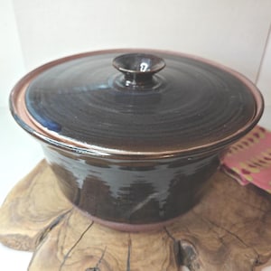 Może przedstawiać: Ręcznie robione, ciemnobrązowe ceramiczne naczynie do zapiekania z pasującą pokrywką. Naczynie ma zaokrąglony kształt z lekko rozszerzonym brzegiem i małym, zaokrąglonym uchwytem na pokrywce. Naczynie stoi na drewnianej powierzchni.