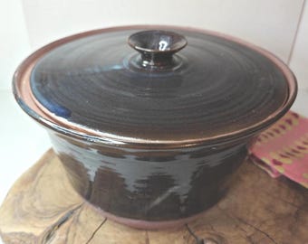 Può includere: Una pirofila in ceramica marrone scuro fatta a mano con coperchio abbinato. Il piatto ha una forma arrotondata con un bordo leggermente svasato e una piccola maniglia arrotondata sul coperchio. Il piatto è appoggiato su una superficie di legno.
