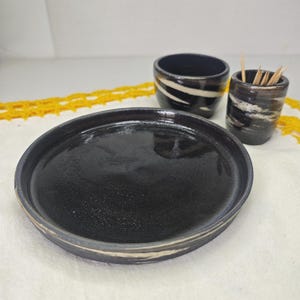 Puede incluir: Un plato de cerámica negro y dos tazas a juego con diseños de rayas blancas y negras. Una taza contiene palillos de dientes. Los artículos están sobre una superficie blanca con un ribete de encaje amarillo.