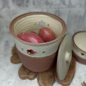 May include: A handmade ceramic potato keeper with a lid. The keeper is two-toned, with a light beige upper section and a reddish-brown lower section. It has star-shaped cutouts and contains several red potatoes. The keeper sits on a wooden trivet.