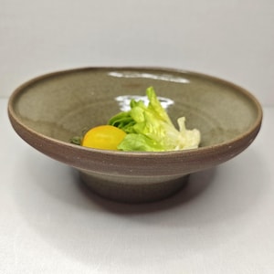 Può includere: Una ciotola da portata in ceramica fatta a mano con un design ampio e poco profondo. La ciotola ha un interno verde chiaro e un bordo e una base marrone scuro. Contiene un pomodoro giallo e lattuga verde. La ciotola è adatta per insalate.