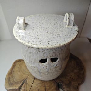 Puede incluir: Un recipiente de cerámica blanca moteada con tapa y dos asas de bucle. El recipiente tiene dos aberturas en forma de ojo y un efecto de esmalte goteado. El recipiente está sobre una base de madera.