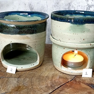 Op de afbeelding: Twee keramische oliebranders met een gespikkelde, gebroken witte basis en een donkerblauwe rand. Eén brander heeft een turquoise interieur en een kaars. De andere heeft een turquoise interieur en een donkergroene band. Beide hebben een ovale opening.