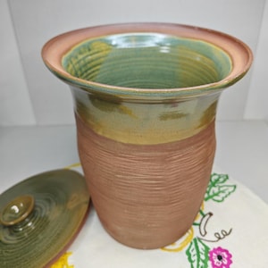 May include: A handmade ceramic jar with a lid. The jar has a textured, reddish-brown body, a yellow band, and a green interior. The rim is a light pink color. The lid is green with a small knob. The jar is sitting on a white cloth.