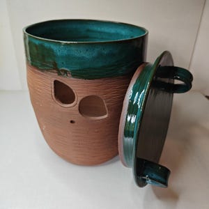 Puede incluir: Un recipiente de cerámica hecho a mano con borde y tapa esmaltados en color turquesa. La parte inferior no está esmaltada, revelando una arcilla marrón rojiza texturizada. El recipiente tiene dos recortes ovalados y un pequeño agujero, con una tapa turquesa a juego.