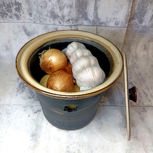 May include: A ceramic garlic and onion keeper with a dark blue-gray exterior and a tan rim. The container holds several onions and a mesh bag of garlic. The lid is off to the side, revealing the contents. A small hole is visible on the side.