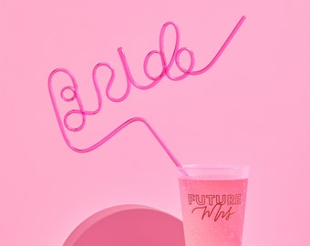 May include: Pink plastic cup with the text "FUTURE Mrs." printed on it. A pink bendy straw is shaped to spell "bride" and is inserted into the cup. The cup is sitting on a red cylinder on a pink half-circle.