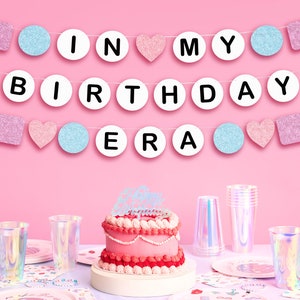 May include: Pink and blue birthday banner with the words "In My Birthday Era" and pink and blue glitter shapes. A pink and white cake with a pink frosting and a topper that says "Happy Birthday" is in front of the banner.