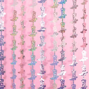 May include: A silver and iridescent garland of cowboy boots with stars. The garland is hanging against a pink background.